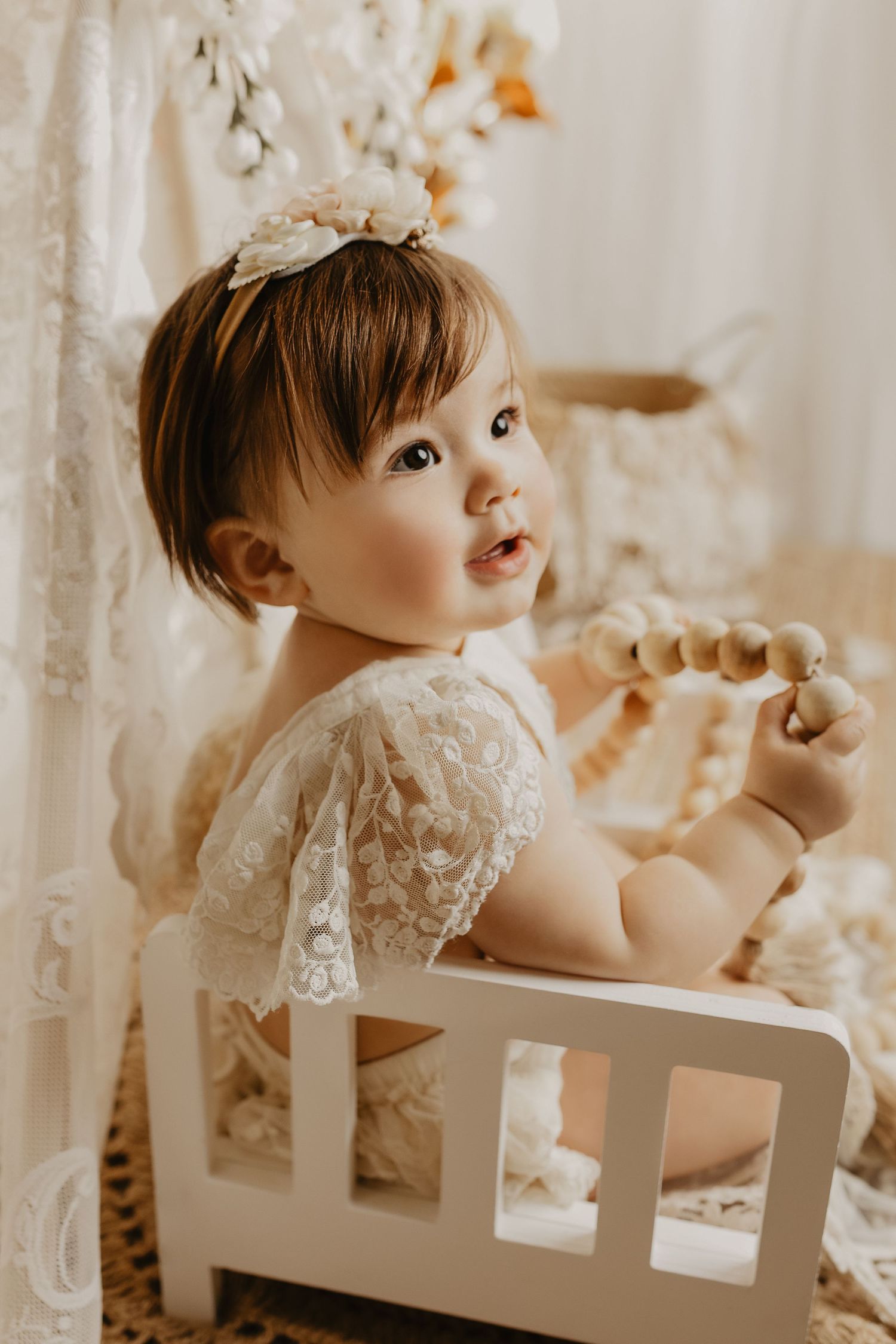 Adorable baby girl in white lace dress and floral headband sitting in a miniature white crib holding wooden beads.