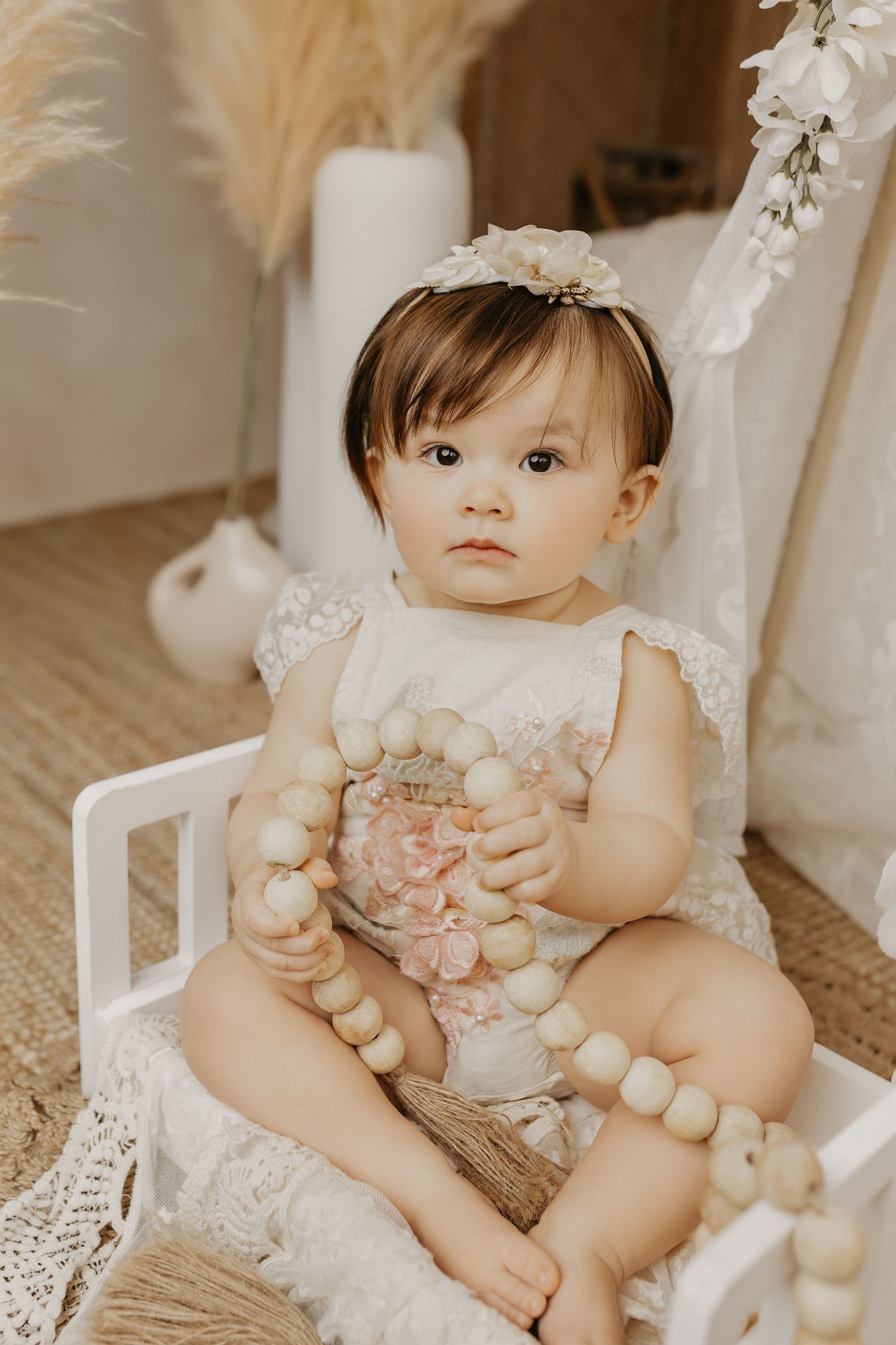 Adorable baby girl in floral romper holds wooden bead garland in boho studio setting.
