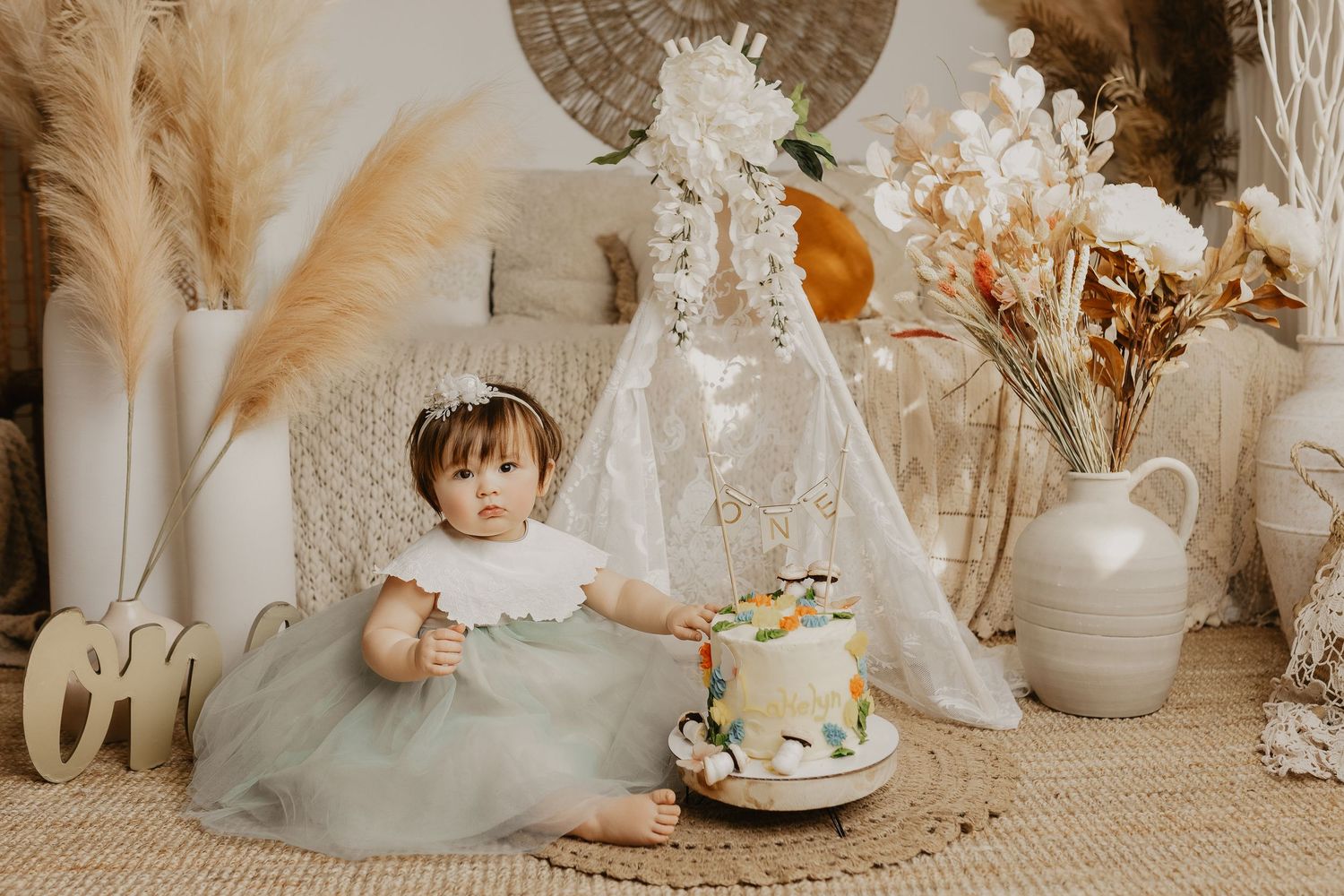 Baby girl in green tutu sits beside smash cake surrounded by boho decor with pampas grass and white teepee.