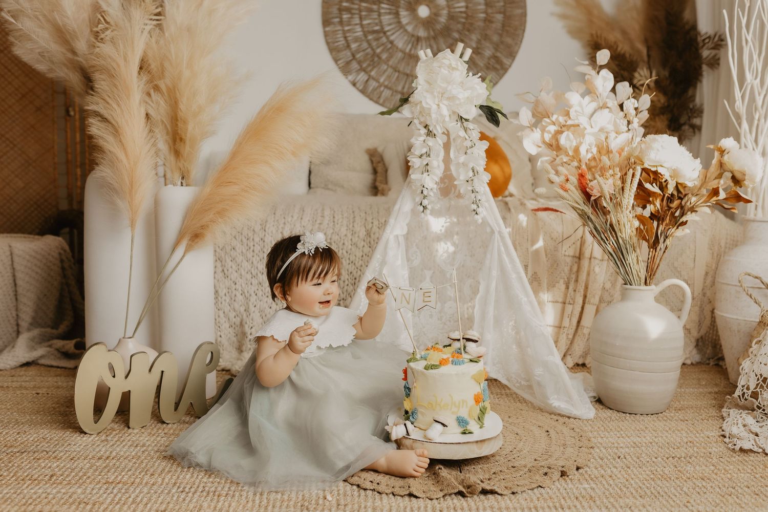Baby girl in white dress touches first birthday cake beside boho teepee with pampas grass and dried florals.