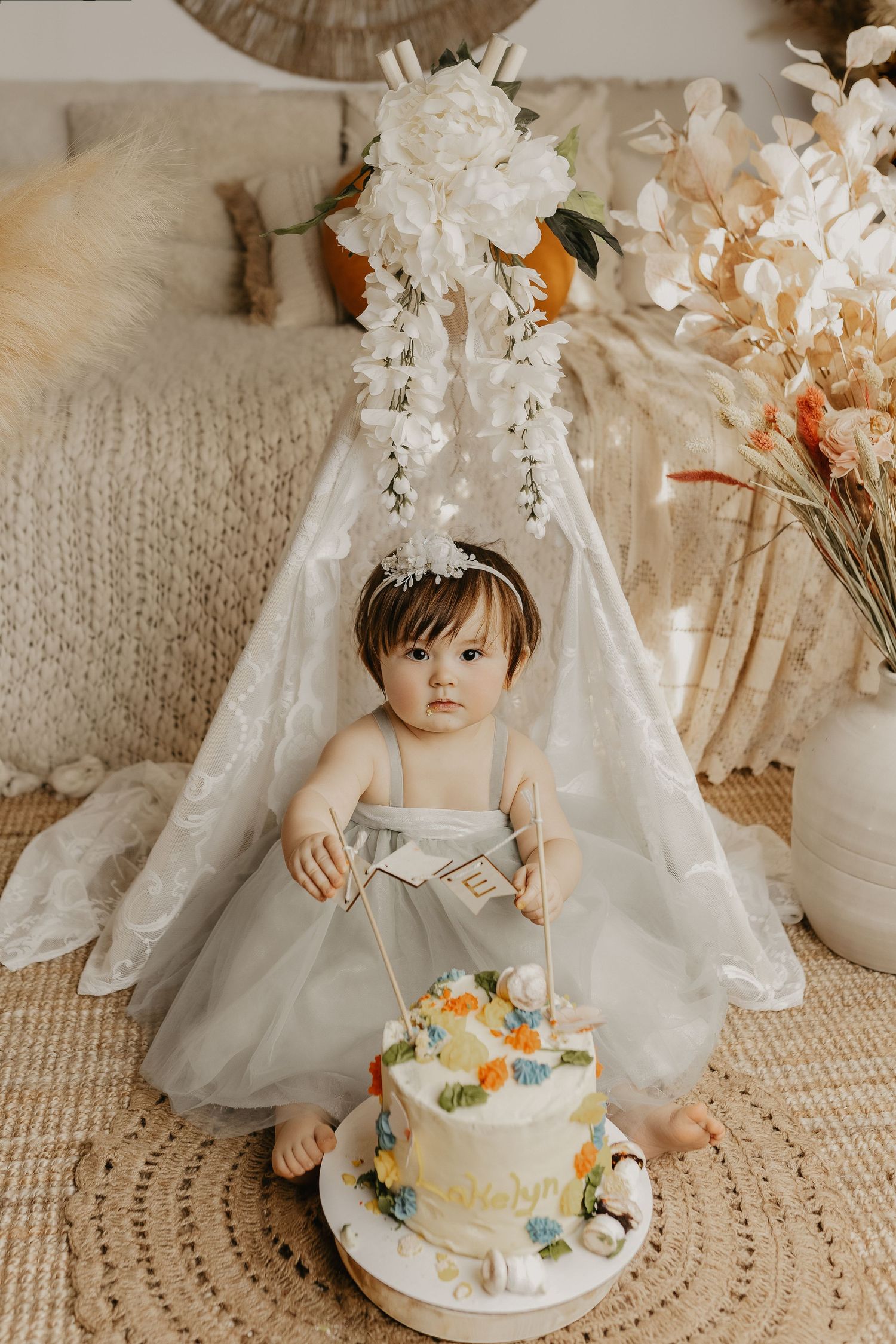 Sweet baby girl in gray tulle dress with floral headband sitting beside a decorated first birthday smash cake.