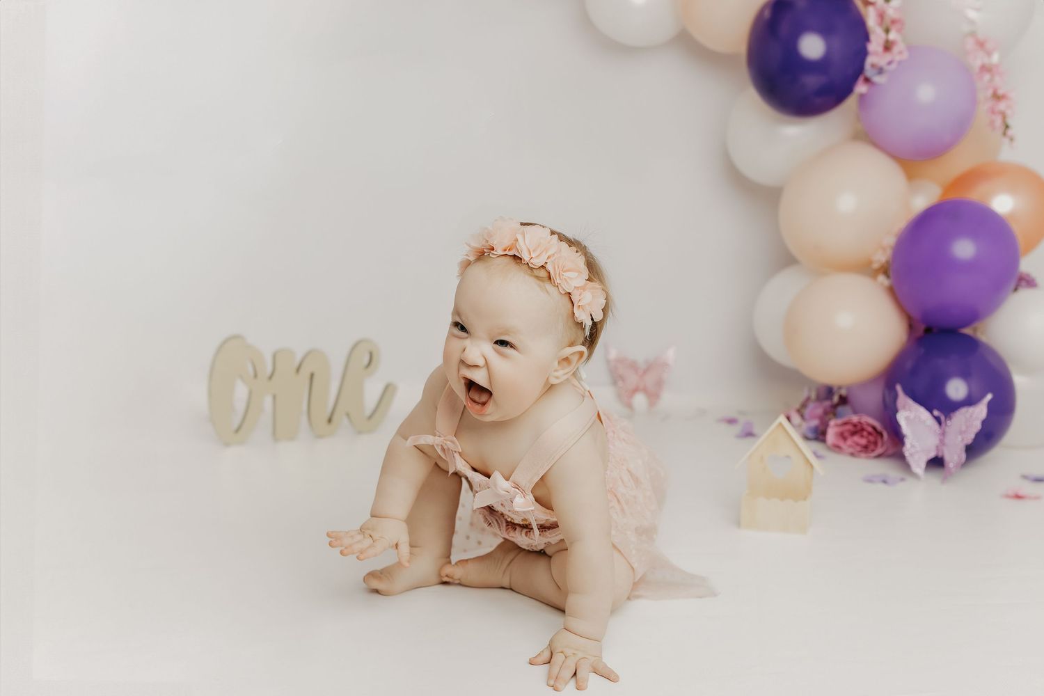 A joyful baby girl in a pink outfit crawls near a balloon arch and \"one\" sign at a first birthday smash cake session.