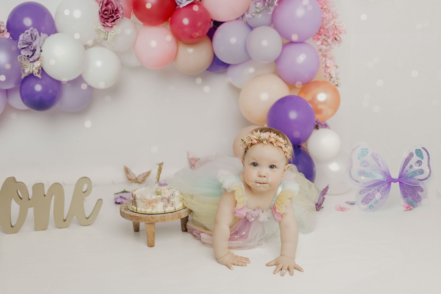 Baby girl in tutu crawls near first birthday cake surrounded by colorful balloons and butterfly decorations.