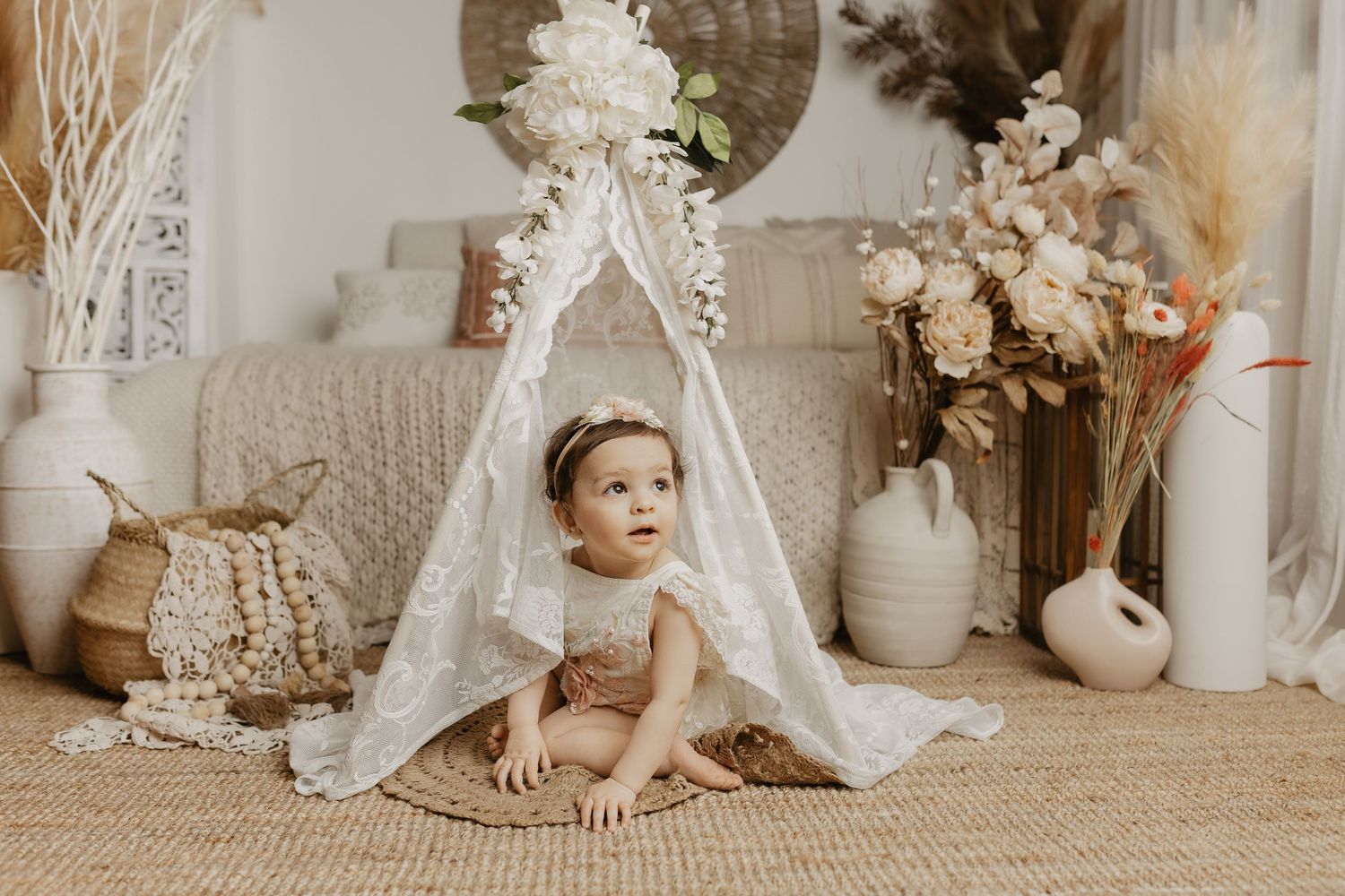 A baby girl sits on a woven rug inside a lace teepee surrounded by boho floral decor and dried pampas grass.