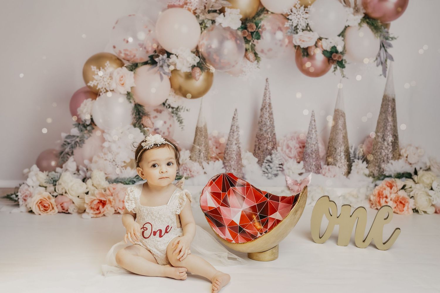 Baby girl in white One onesie sits before pink balloon arch with flowers and decorative trees for first birthday.