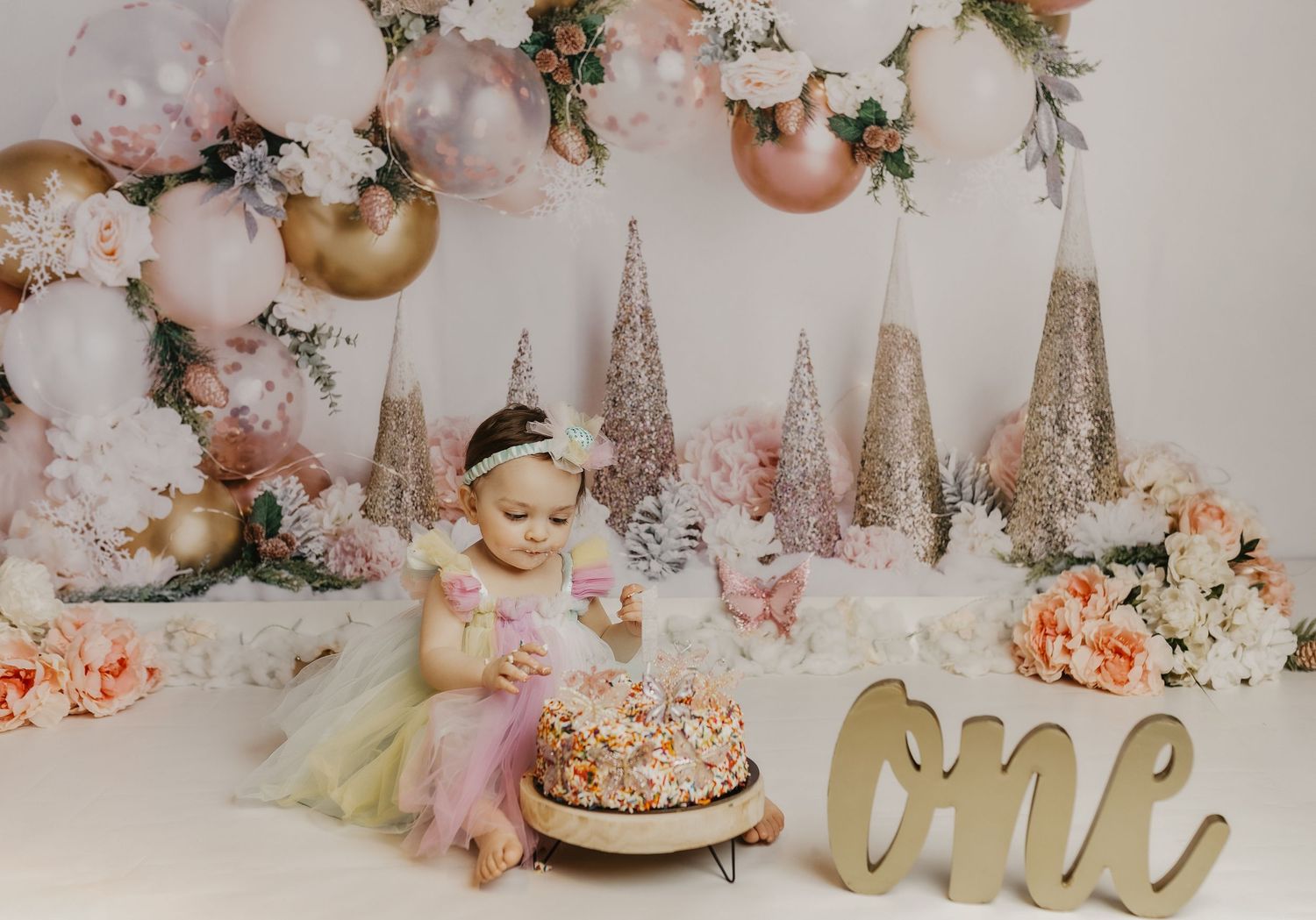 Baby girl in tutu sits before a cake at her first birthday party with pink balloons and floral decorations.