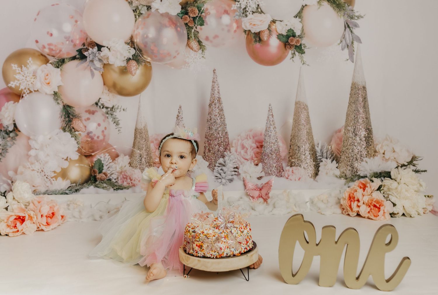 Baby girl in tutu celebrates first birthday with colorful smash cake, pink floral balloon arch backdrop.