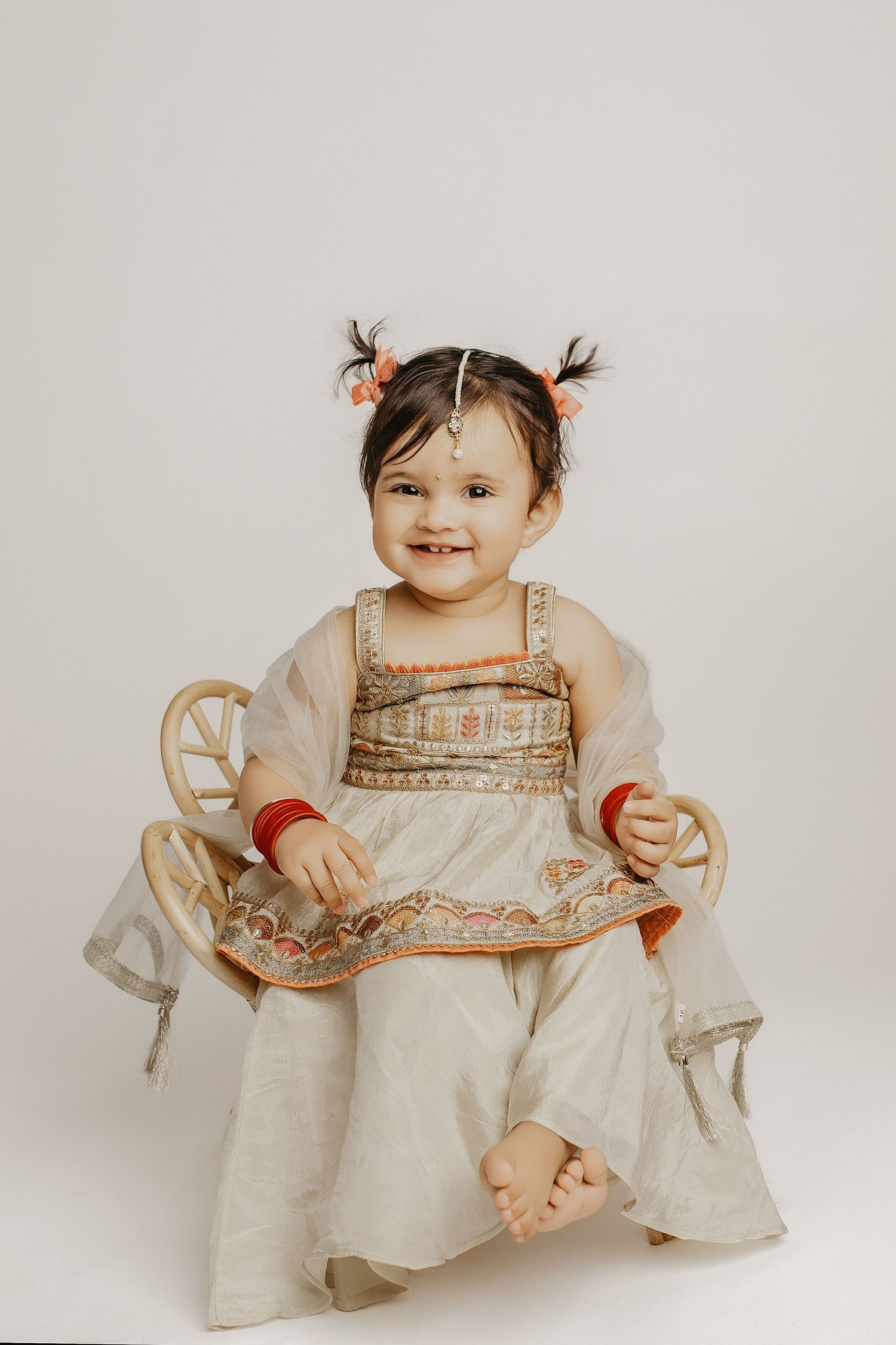 Smiling baby girl in traditional Indian white and gold outfit with pigtails, sitting on decorative chair.
