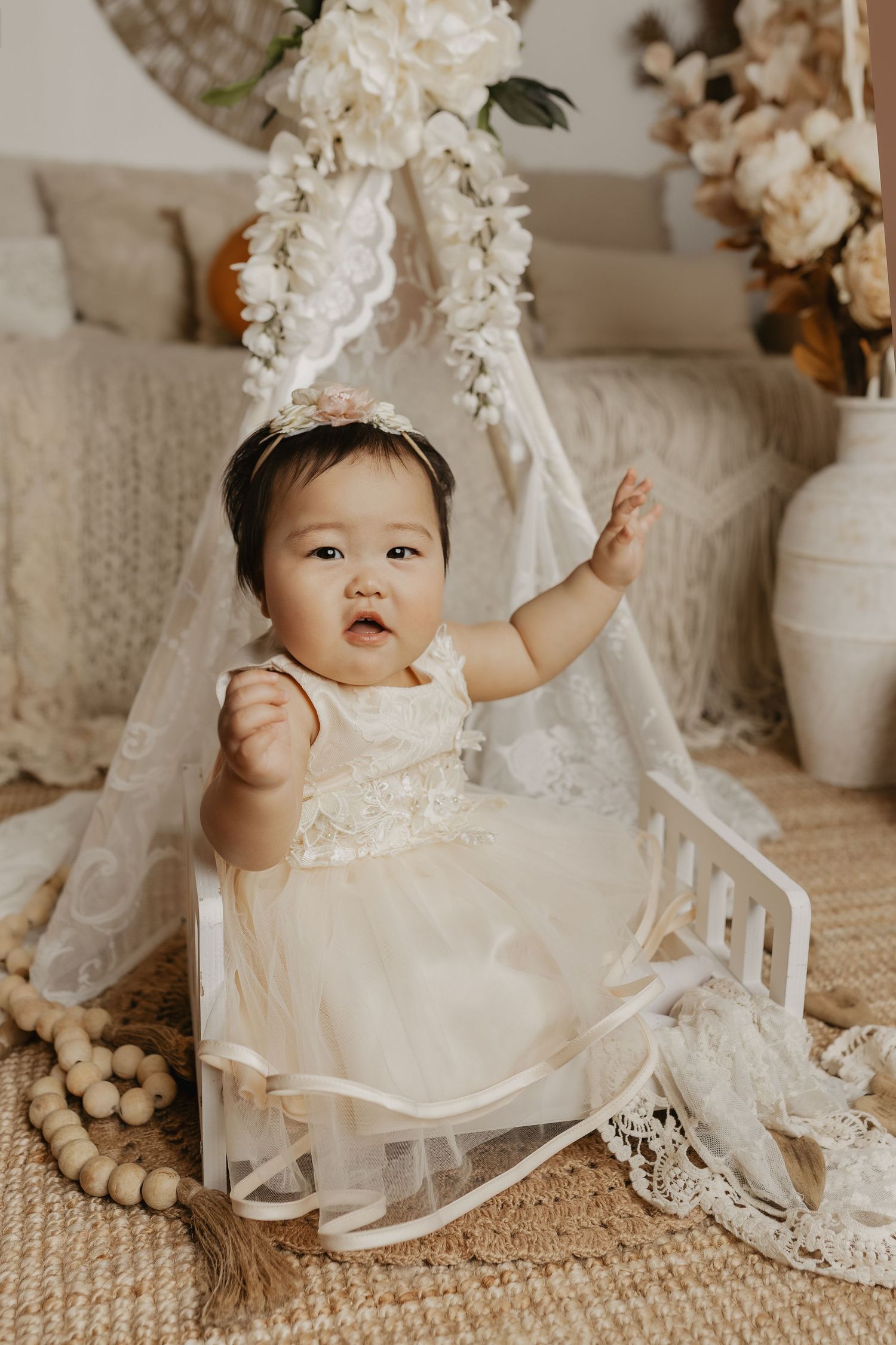 Baby girl in ivory tulle dress and floral crown sits in mini crib with boho floral backdrop.