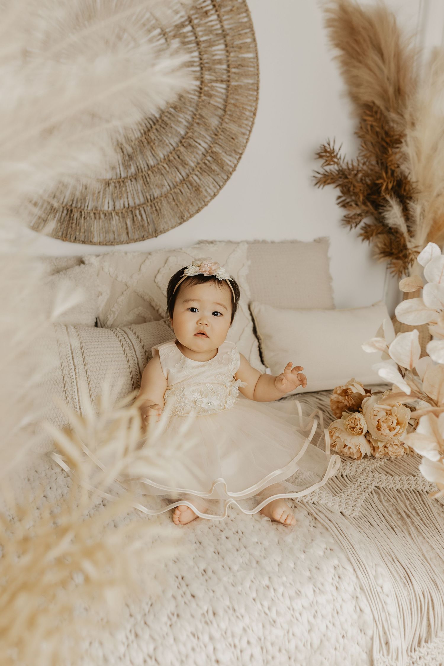 Baby girl in white dress and headband sits on boho-styled bed with pampas grass and wicker decor.