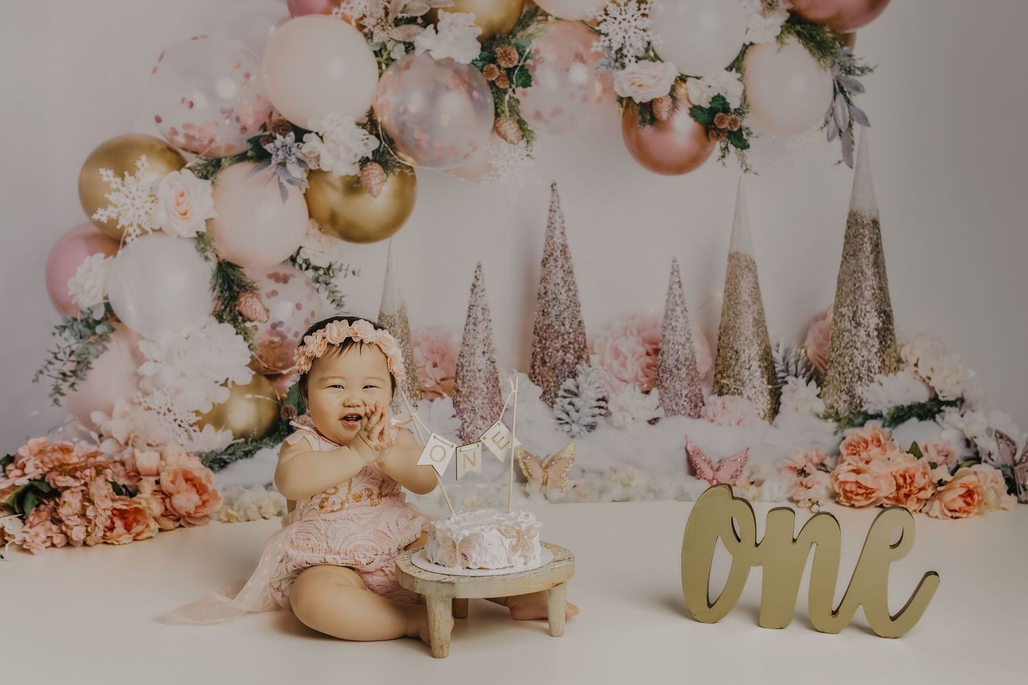 Happy baby girl in pink dress celebrates first birthday with cake smash and balloon arch.