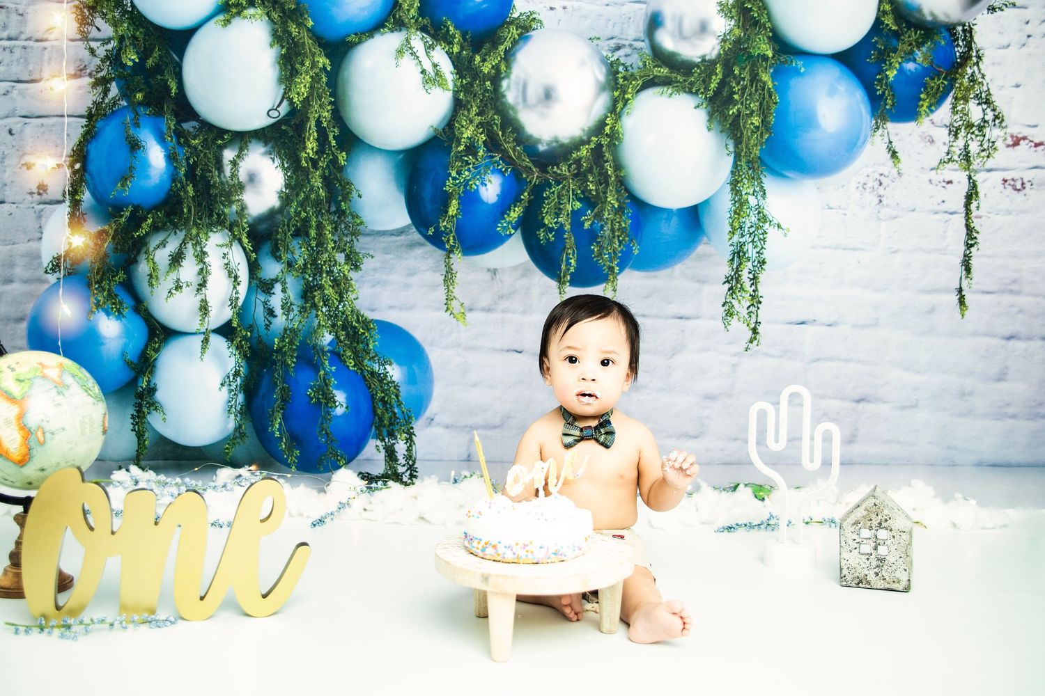 First birthday photo setup with blue balloons, white brick wall, and gold decorations.