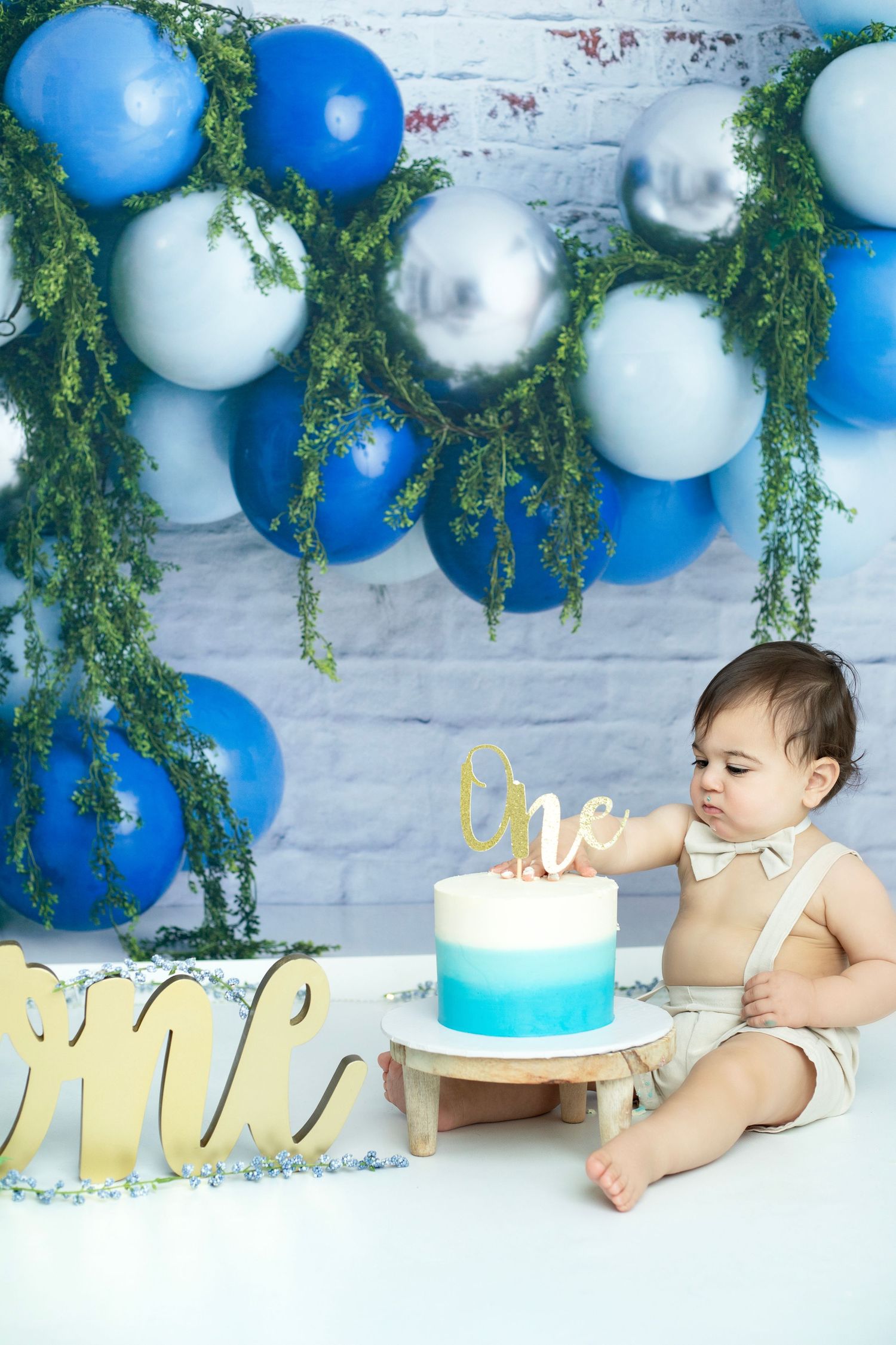 First birthday cake smash setup with blue balloons, greenery, and gold 'one' sign.