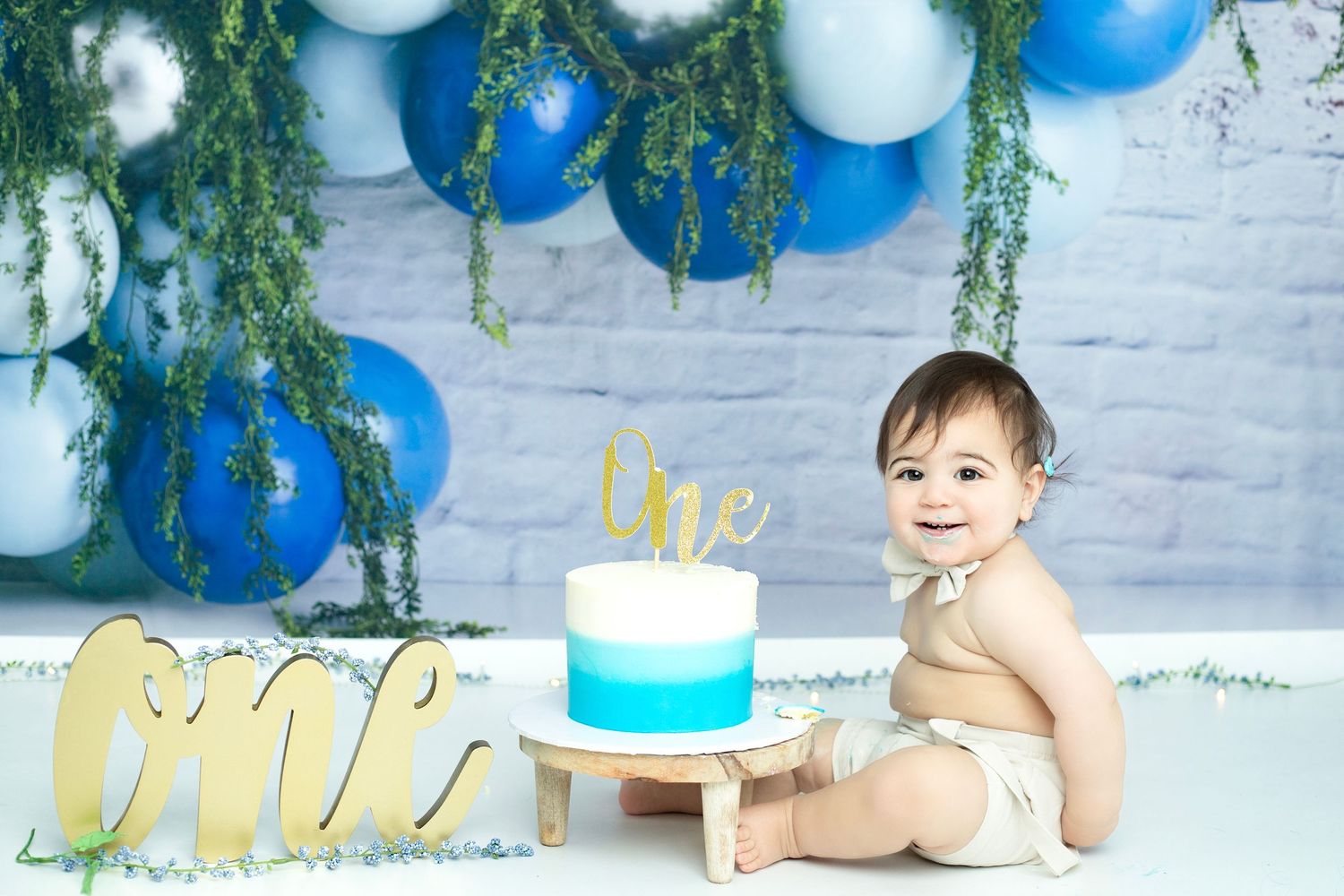 Blue and gold themed first birthday cake smash setup with hanging greenery.