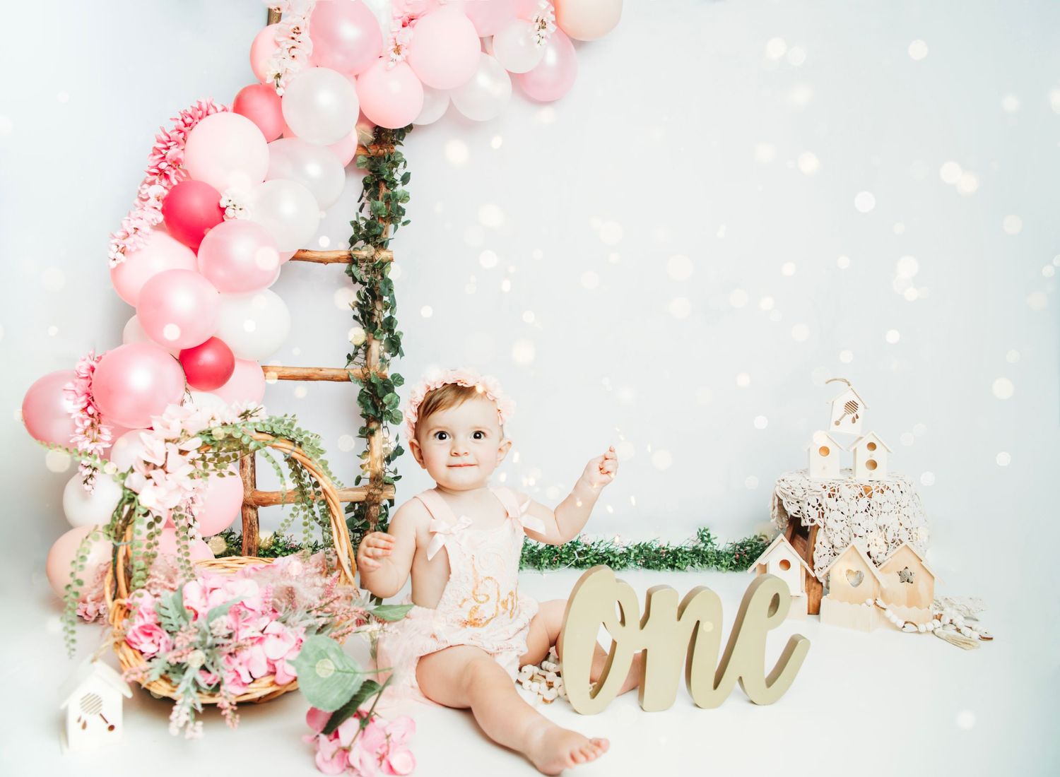Baby photo session setup with pink balloons, floral decorations, and rustic wooden ladder against white backdrop.