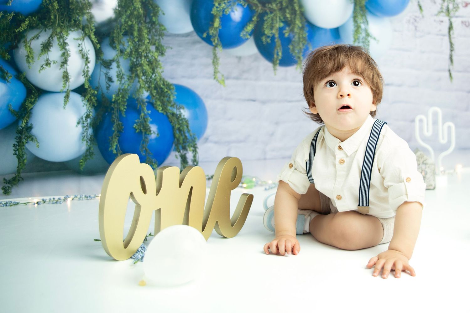 Baby in white shirt and suspenders sits against blue and white balloon backdrop with gold 'one' decoration.
