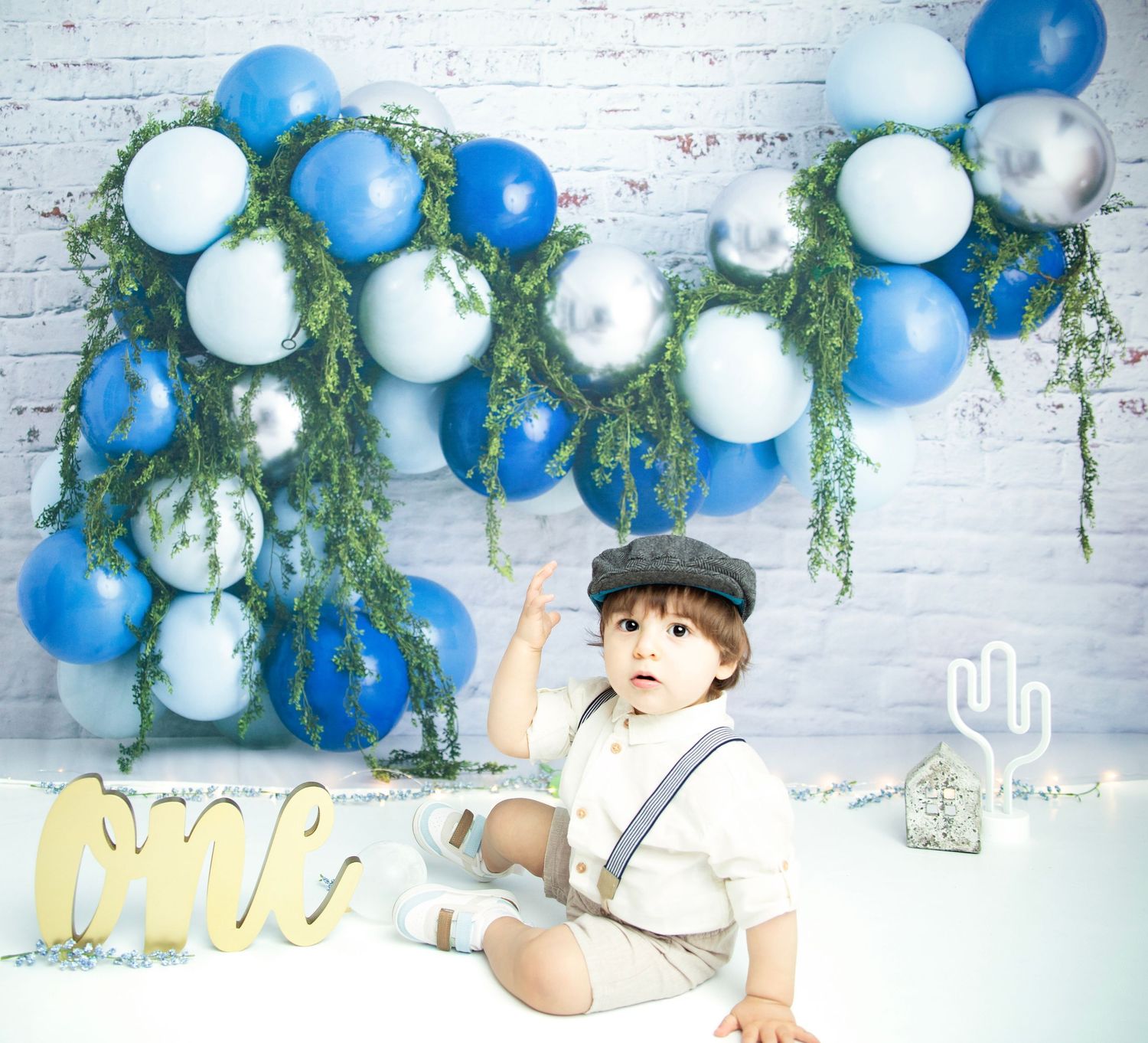 Toddler in vintage outfit poses with blue and white balloons and greenery decorations.