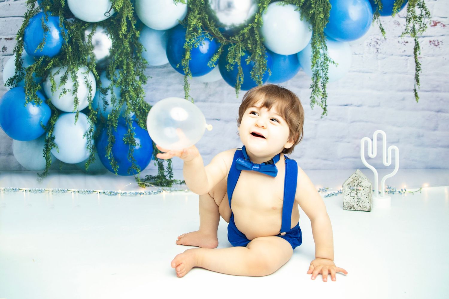 First birthday photo setup with blue and white balloons, greenery, and gold 'one' sign.