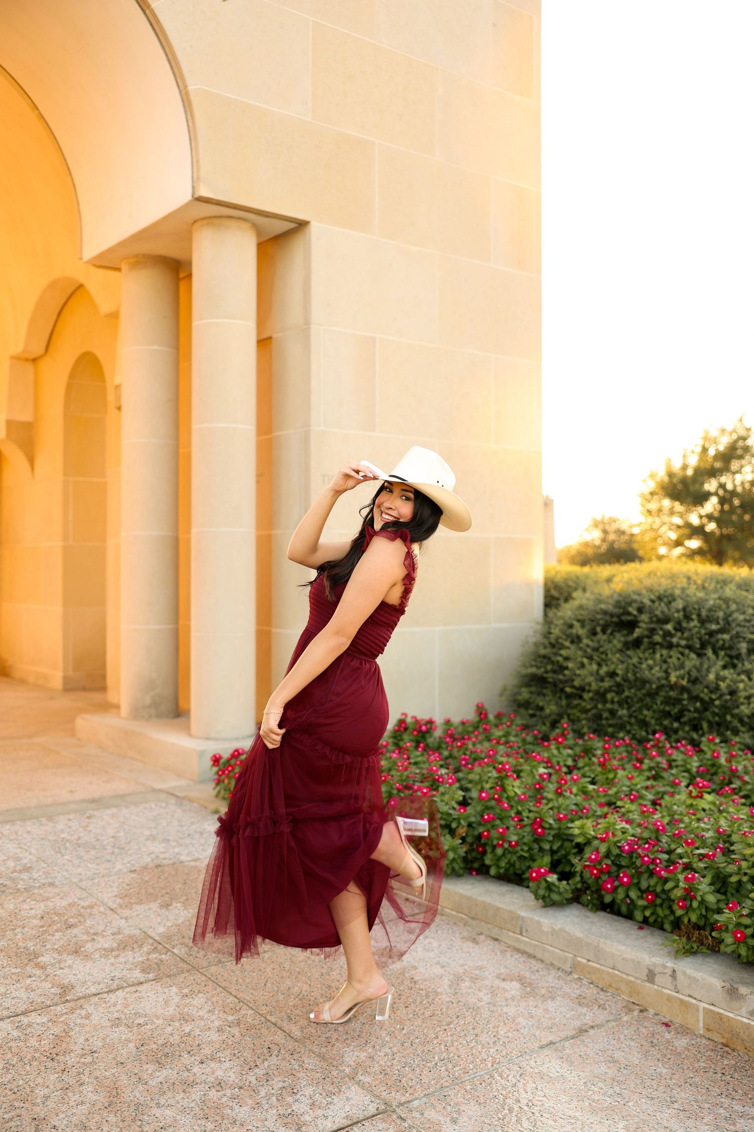Dancer in flowing burgundy dress performs graceful movements among arched columns at golden hour.