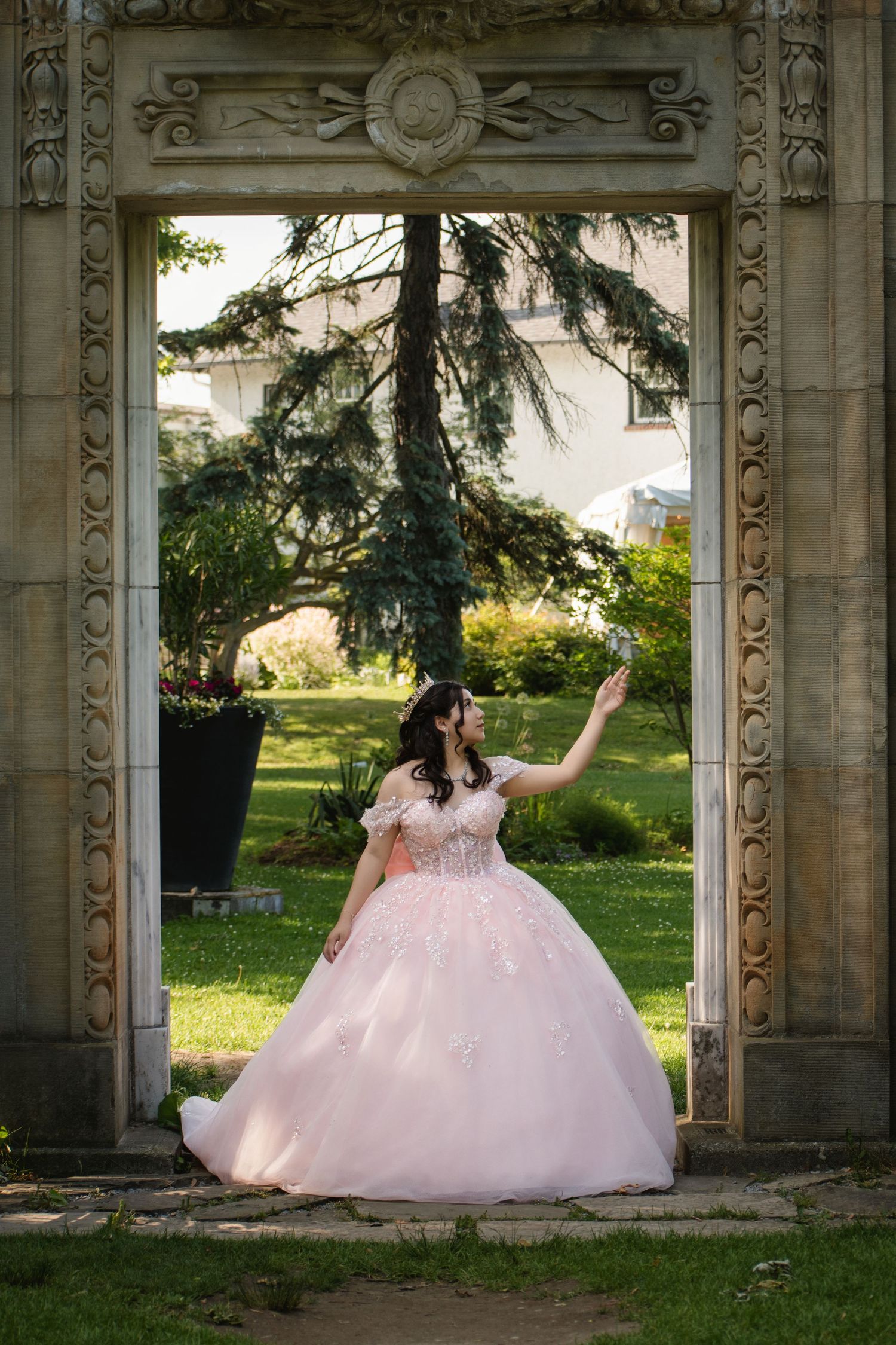 Young woman in pink ball gown posing in ornate stone archway with lush garden background. Toronto GTA