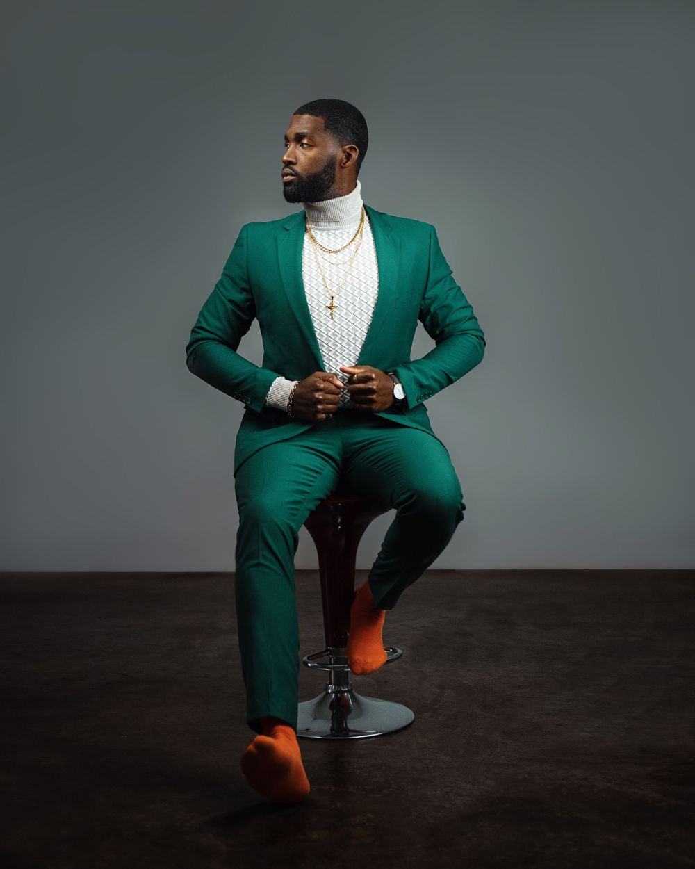 Fashion portrait of model in emerald green suit and white shirt seated on stool against dark background.