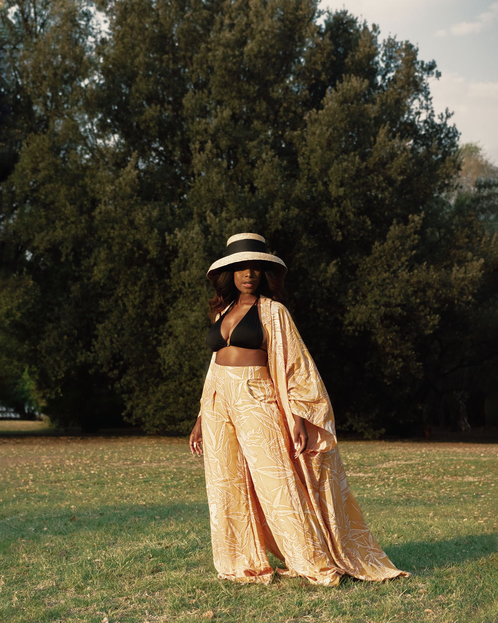 A figure in a sun hat and flowy beige outfit poses elegantly against a backdrop of tall trees in a park setting.