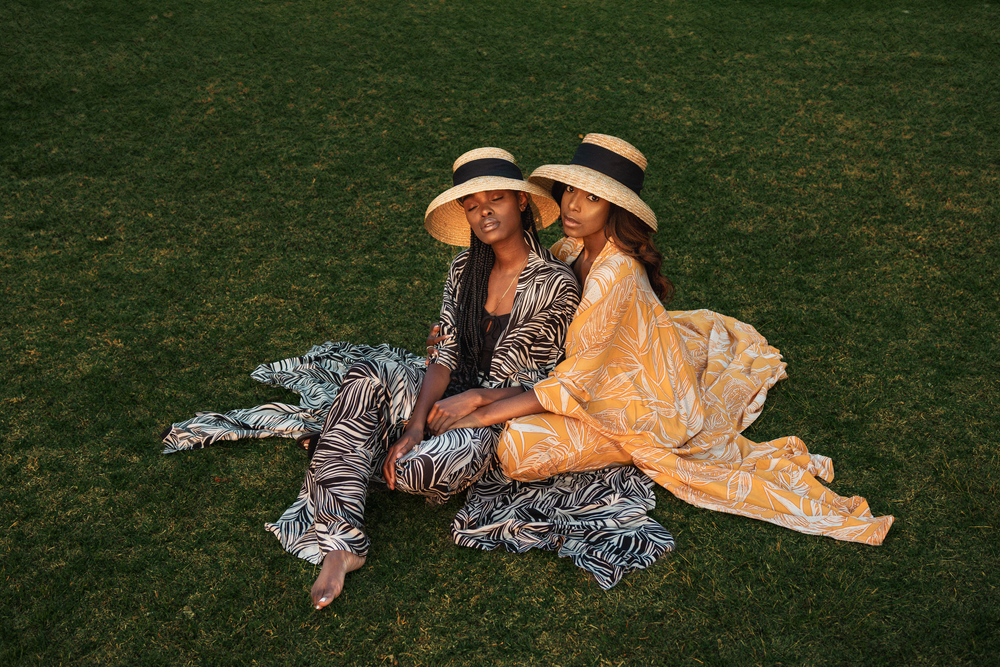 Two people in matching sun hats lounge on grass wearing coordinated flowing outfits in beige and patterned prints.