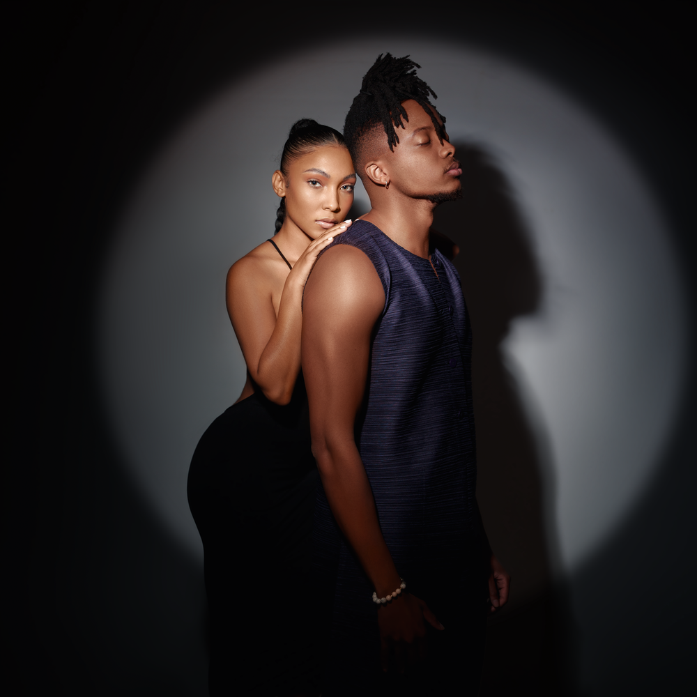 A dramatic spotlight portrait of two models posed back-to-back in formal attire.