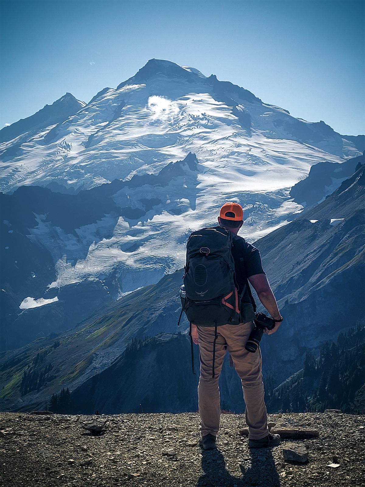A hiker overlooks a snow-covered mountain peak on a clear blue day.