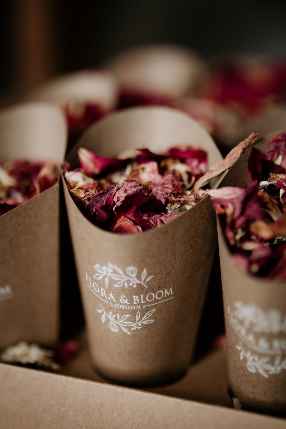 Brown paper cones decorated with white floral designs hold dried rose petals in a romantic wedding detail shot.