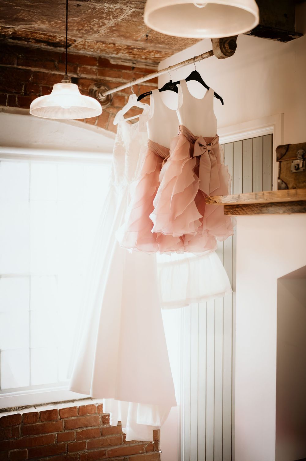 Soft pink tulle dresses hanging from a rustic ceiling with exposed beams and brick walls in a vintage-style room.