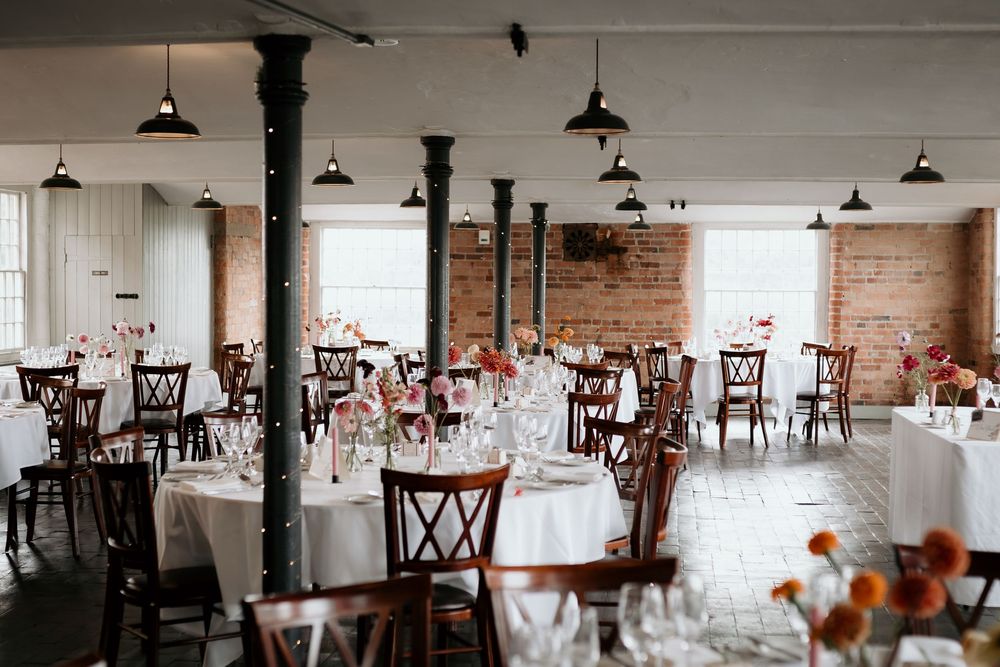 A rustic wedding reception venue with exposed brick walls, white tablecloths, and elegant wooden cross-back chairs.