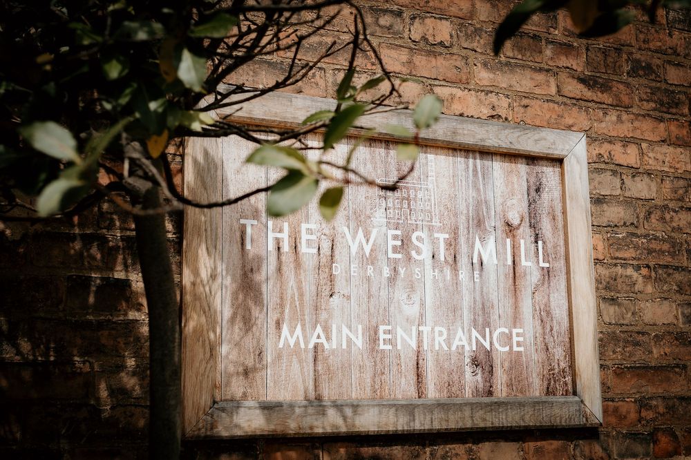 A weathered wooden sign reading 'The West Mill Main Entrance' mounted on a brick wall with green foliage.