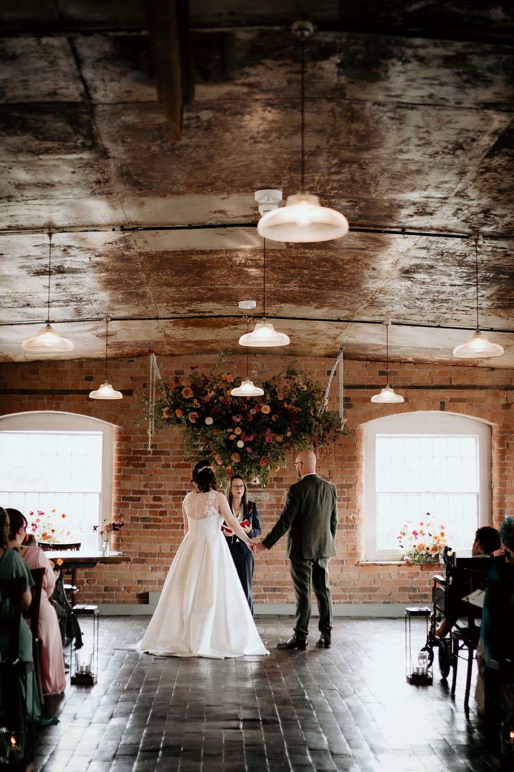 A wedding ceremony takes place in a rustic brick venue with exposed wooden beams and pendant lights overhead.