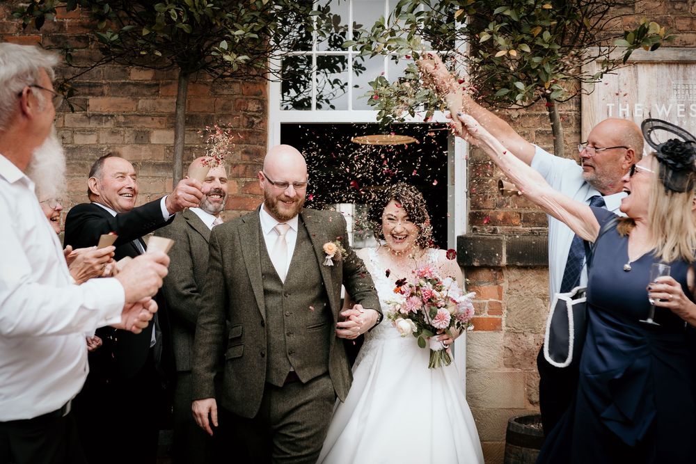 Wedding guests celebrate with bubbles as newlyweds exit their ceremony at a rustic brick venue.