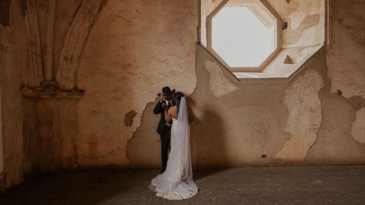 A couple in formal wedding attire descend stone stairs in a rustic architectural setting with weathered walls.