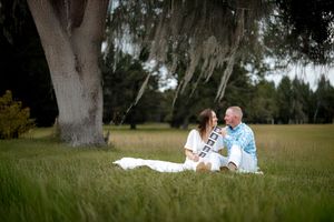 A couple sits together on a blanket under a large oak tree with Spanish moss hanging from its branches.