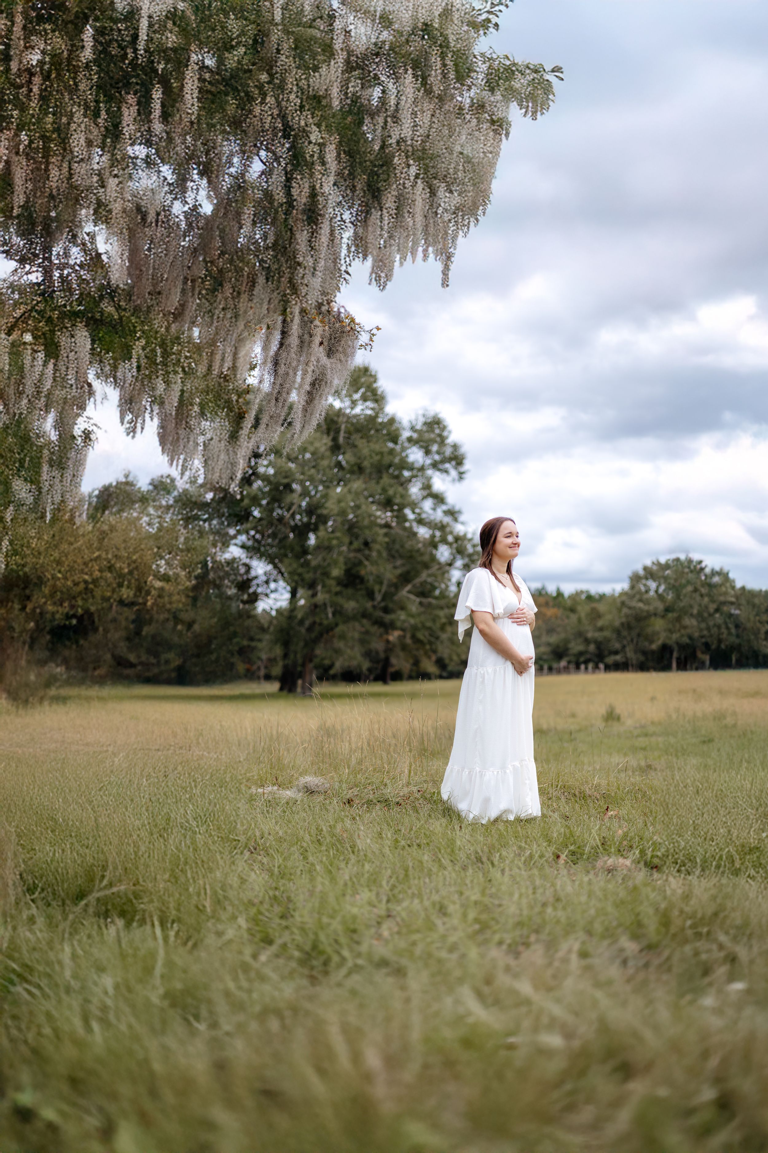 A figure in a white dress stands in a grassy field with Spanish moss hanging from trees in the background.