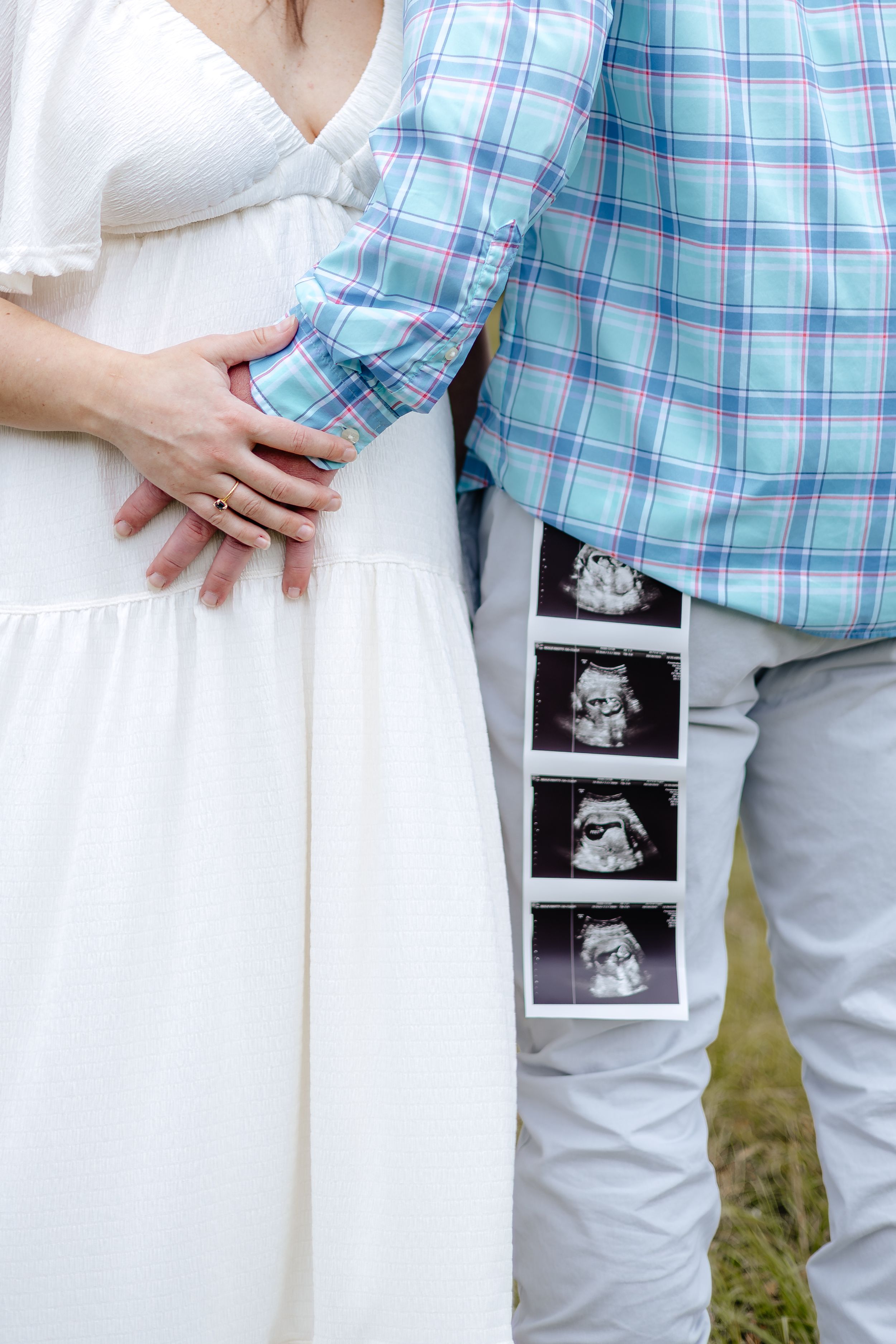Couple in white dress and plaid shirt hold ultrasound photos during pregnancy announcement photoshoot outdoors.