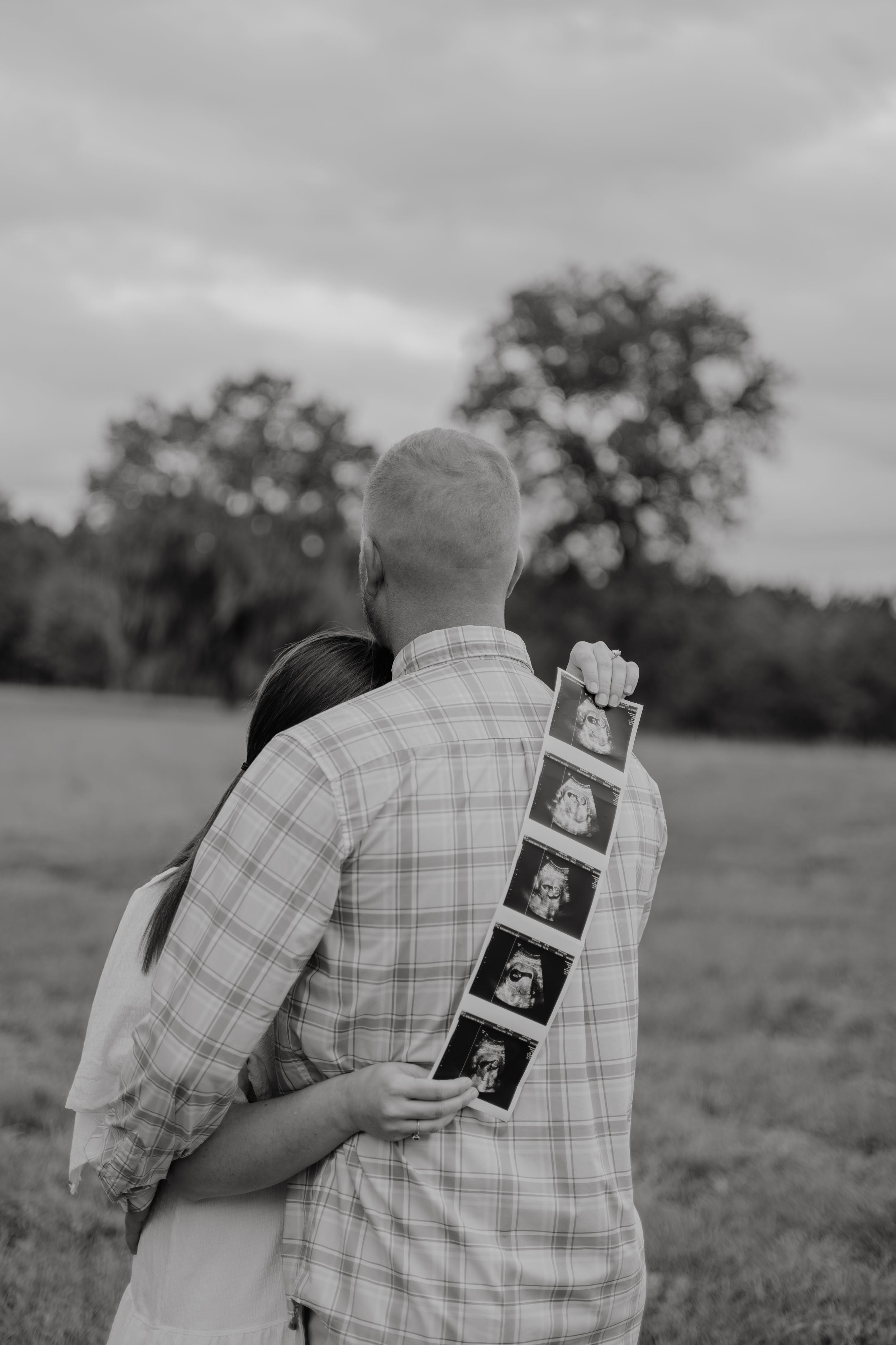 A couple embracing in a field while holding ultrasound photos against a plaid shirt in black and white.
