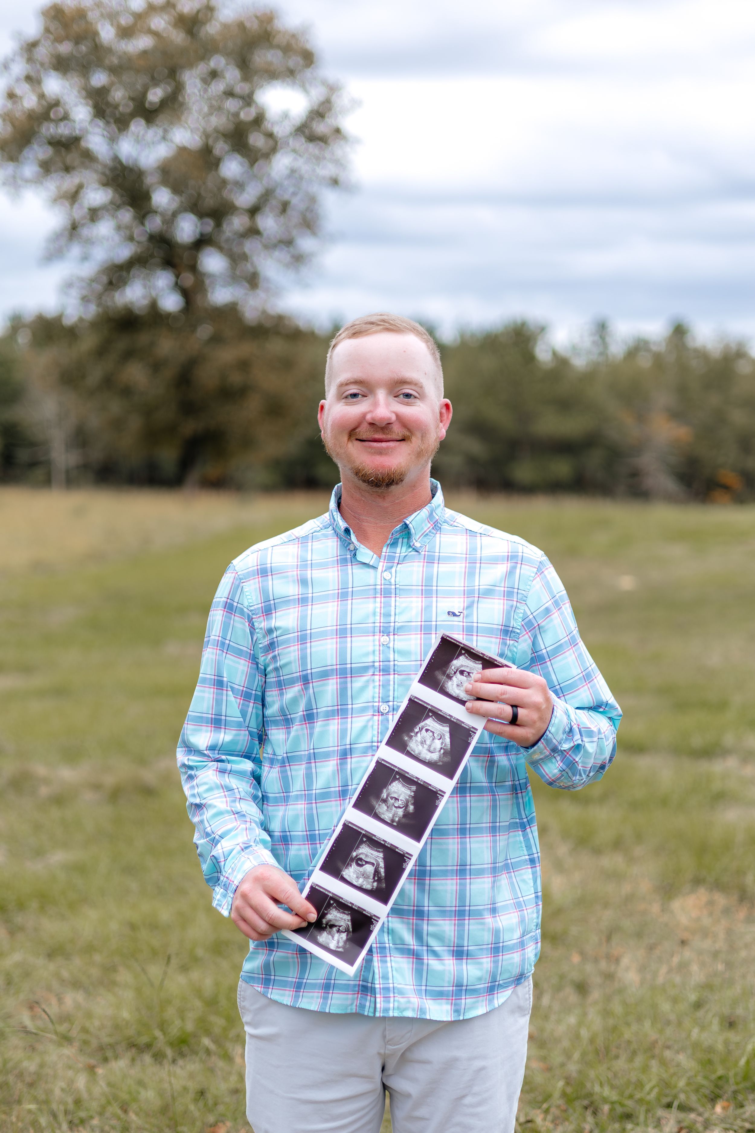 A person in a blue plaid shirt stands in a grassy field holding ultrasound photos while smiling at the camera.