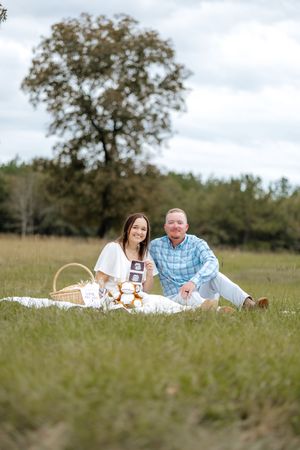 A couple enjoys a romantic picnic on a blanket in a grassy field with a large tree in the background.