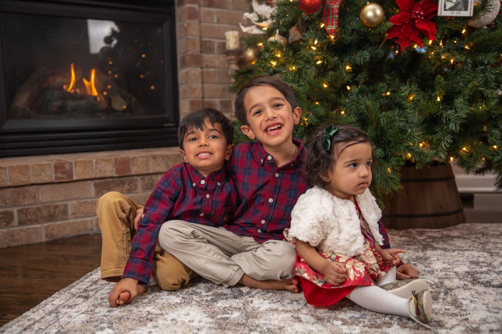 Three children sitting together by decorated Christmas tree and brick fireplace.