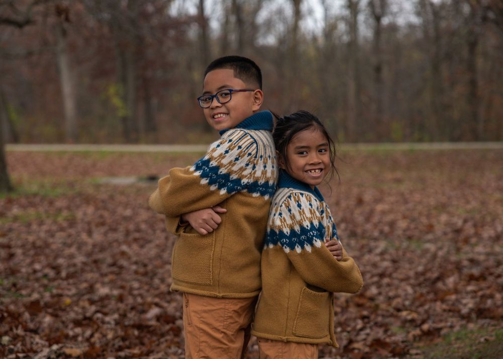 Two children in matching sweaters and brown pants playfully pose in autumn forest.