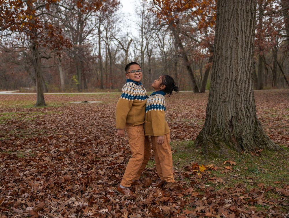 Two children in coordinated outfits embrace while playing in a leaf-covered forest setting.