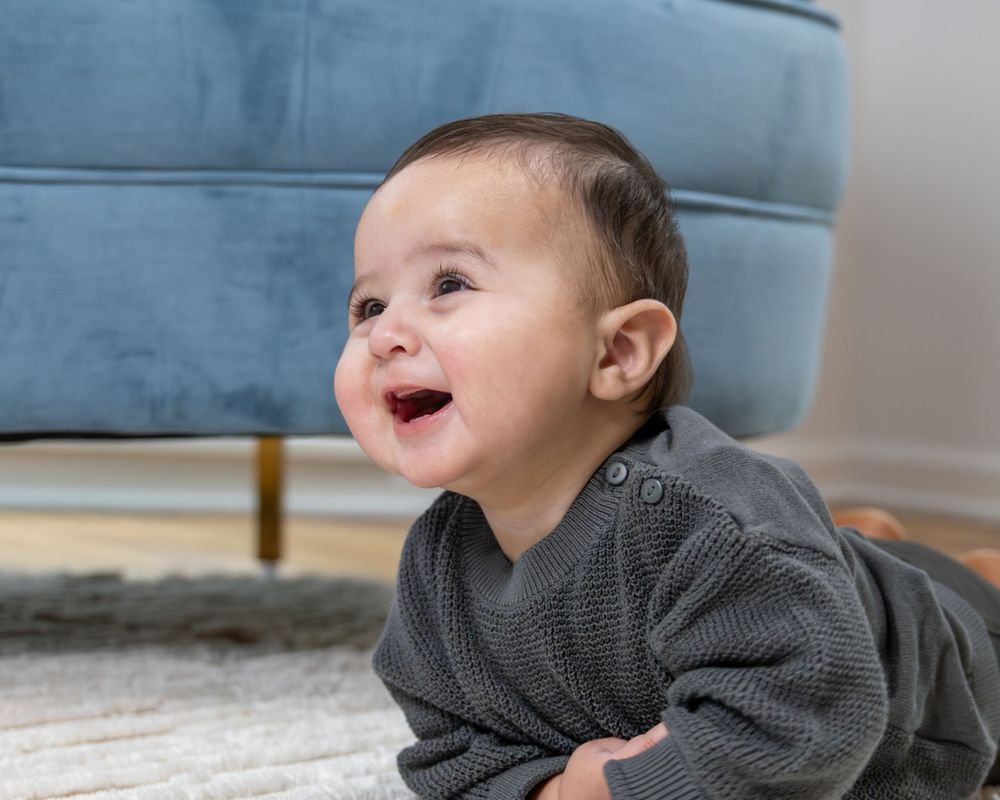 Happy baby in gray sweater lying on carpet smiling with joy and excitement.