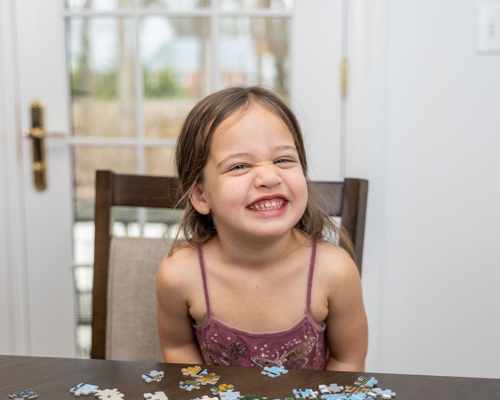 Young child in purple dress smiling with excitement at table with puzzle pieces.