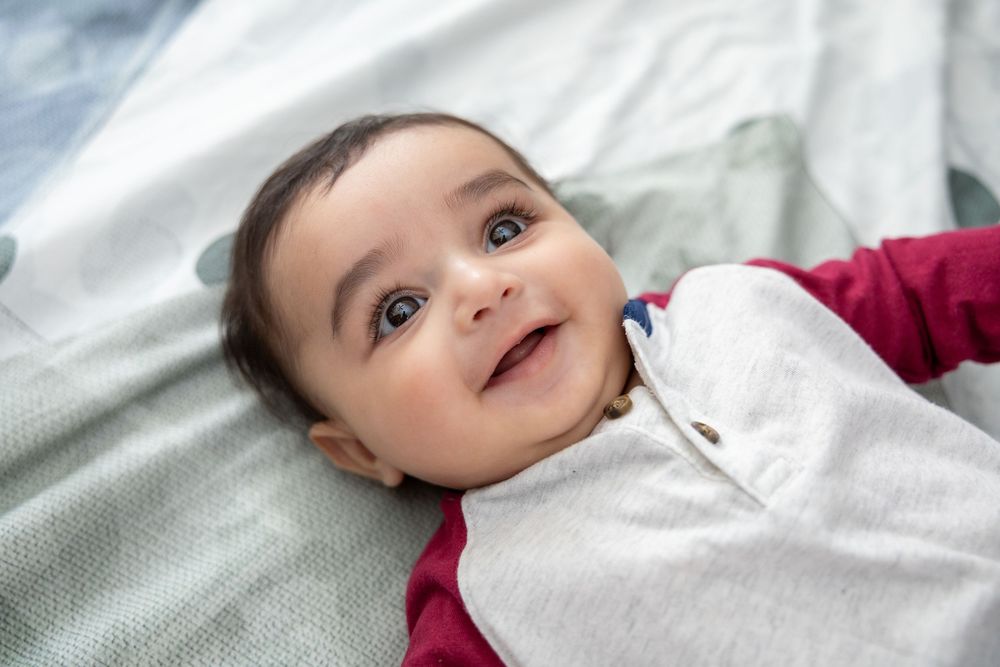 A smiling baby wearing a white and burgundy outfit lies on a light-colored blanket.