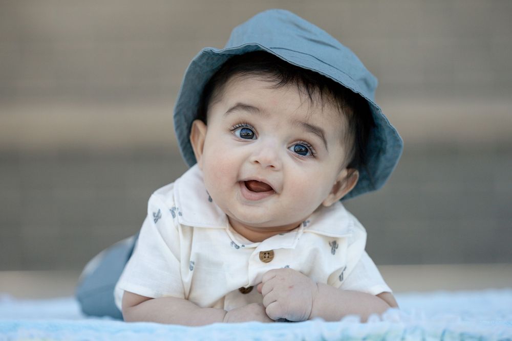 Baby wearing blue hat and white shirt lies on blanket with delighted expression.