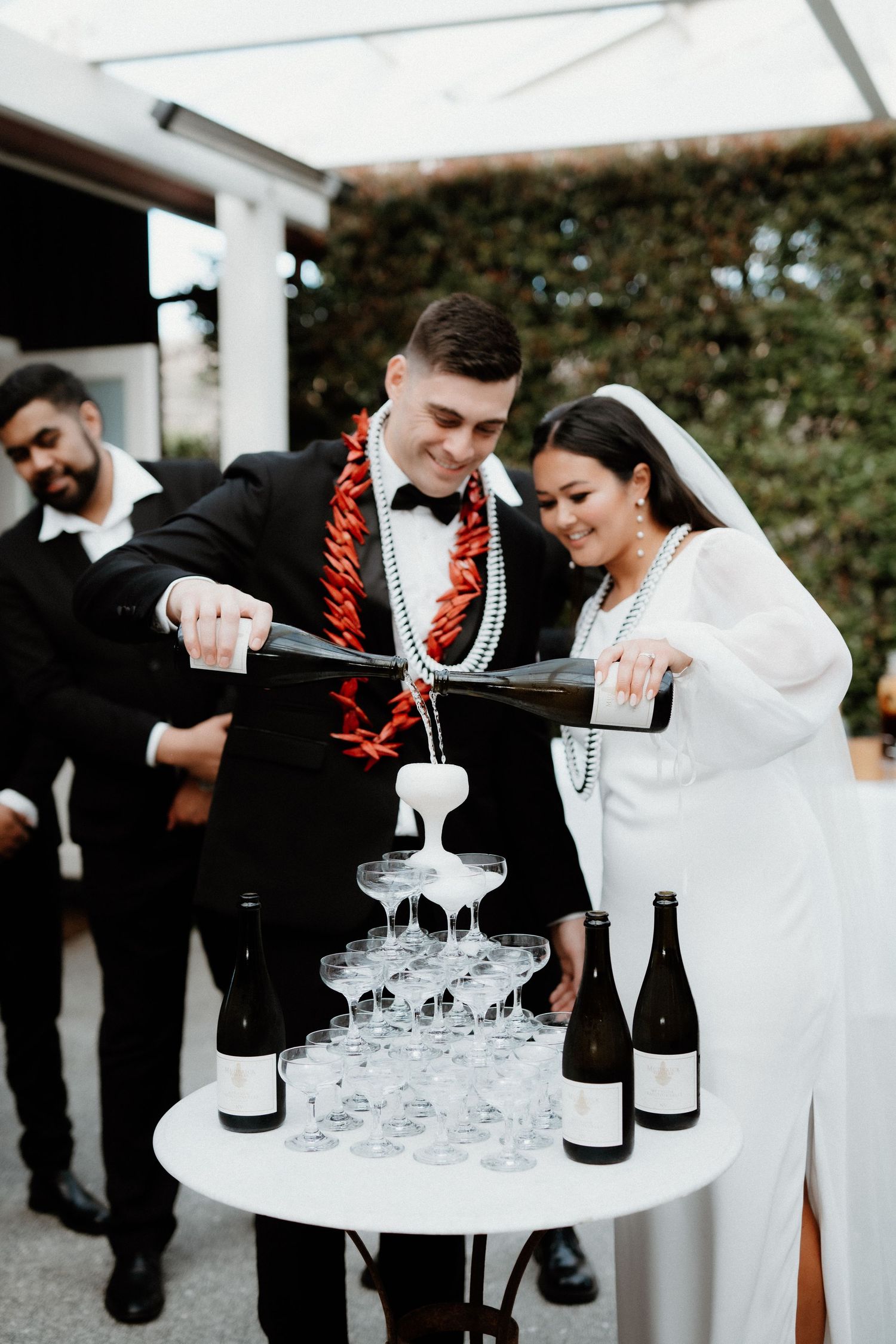 Couple in formal attire pour champagne into a tower of glasses during wedding celebration.